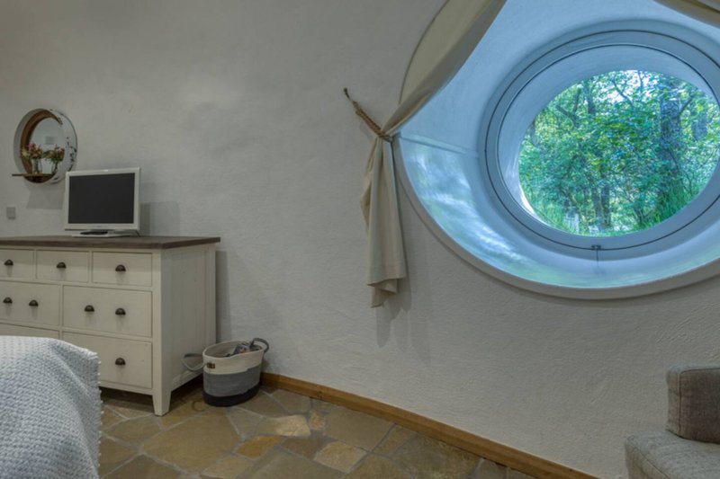 Bedroom with cream dresser, small TV, circular window with forest view, and stone flooring