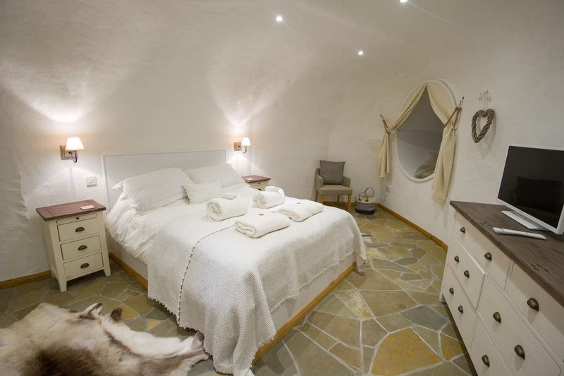 Bedroom with white double bed, wooden nightstands, cream dresser, wall-mounted mirror, and TV