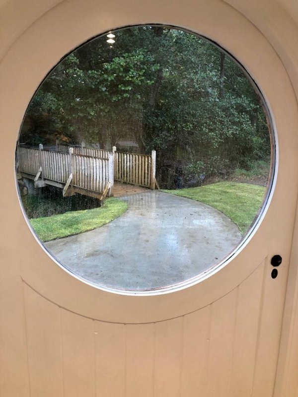 View through round porthole window of driveway, white fence, and tree-lined property