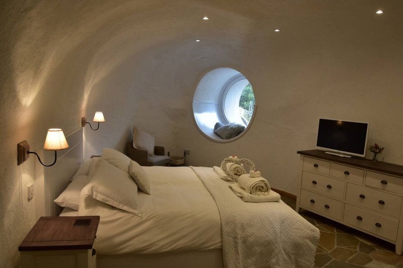 Curved bedroom with arched ceiling, round porthole window, bed with cream bedding, wooden dresser and wall sconces