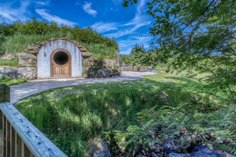 Stone hobbit hole entrance with round wooden door, surrounded by garden and stream