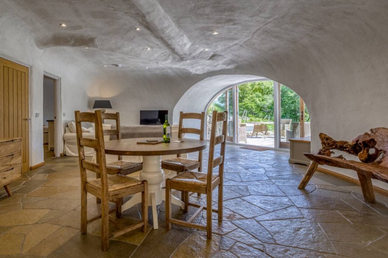 Open-plan living space with dining table, wooden chairs, stone flooring and arched doorway to garden