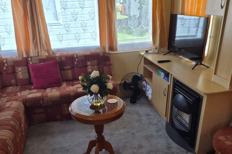 Living room with burgundy patterned sofa, round wooden table with flowers, TV, and windows with orange curtains