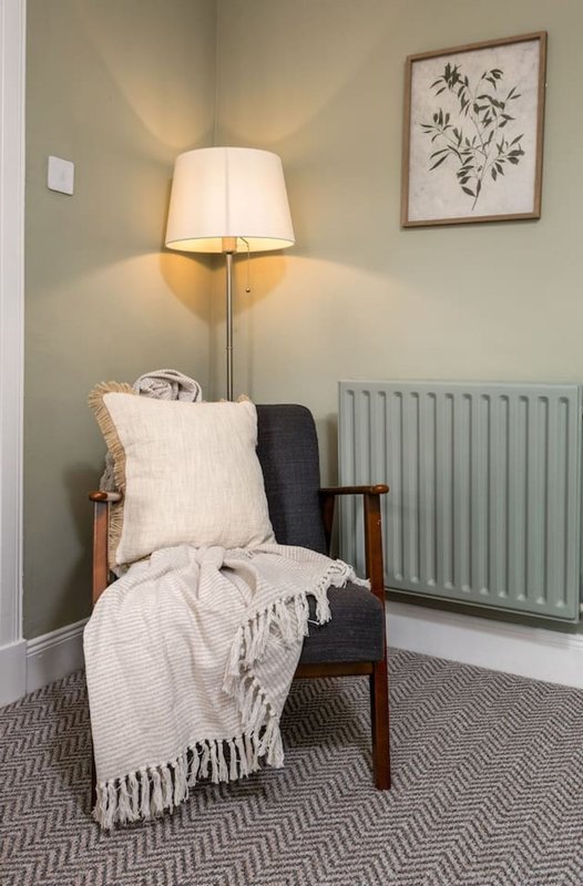 Cozy reading corner with wooden armchair, cream cushion, throw blanket, table lamp and botanical wall art