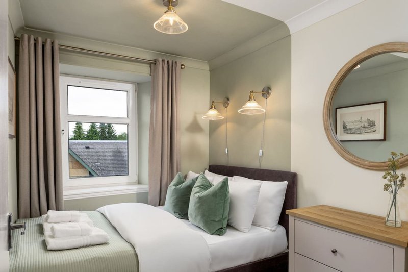 Bright bedroom with double bed, green cushions, wooden dresser, round mirror and pendant lights