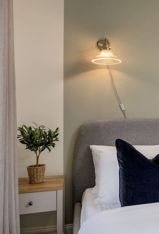 Bedroom corner with gray upholstered headboard, white pillows, black cushion, wall-mounted lamp, and small potted plant on bedside table