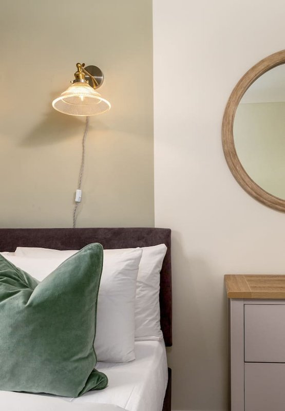 Bedroom with dark headboard, white pillows, green cushion, wall-mounted pendant lamp and round mirror