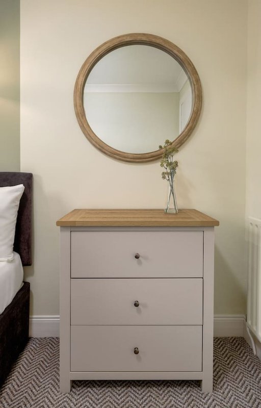 Bedroom corner with round wooden mirror above white chest of drawers and flowers in vase