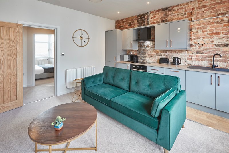 Open-plan kitchen and lounge with teal sofa, brick chimney, wooden dining table