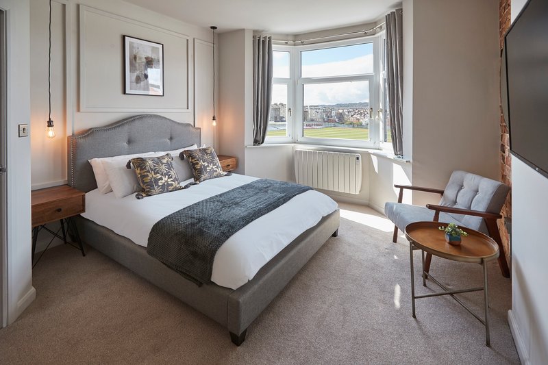 Spacious bedroom with upholstered bed, armchair, curved window with countryside views