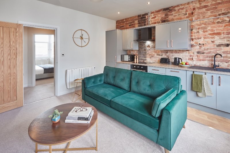 Open-plan living area with teal sofa, kitchenette, exposed brick feature wall and wooden flooring