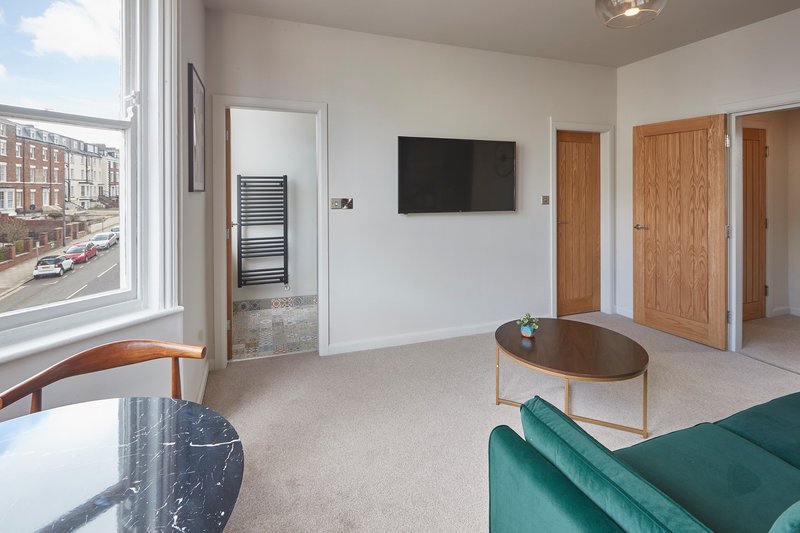 Modern living room with green sofa, wooden coffee table, wall-mounted TV and large windows overlooking street