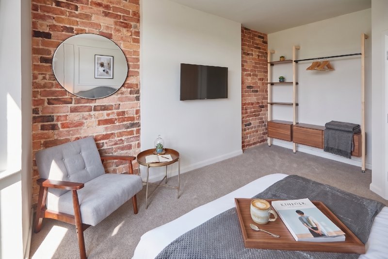 Modern bedroom with exposed brick walls, grey armchair, wooden bed frame and shelving unit
