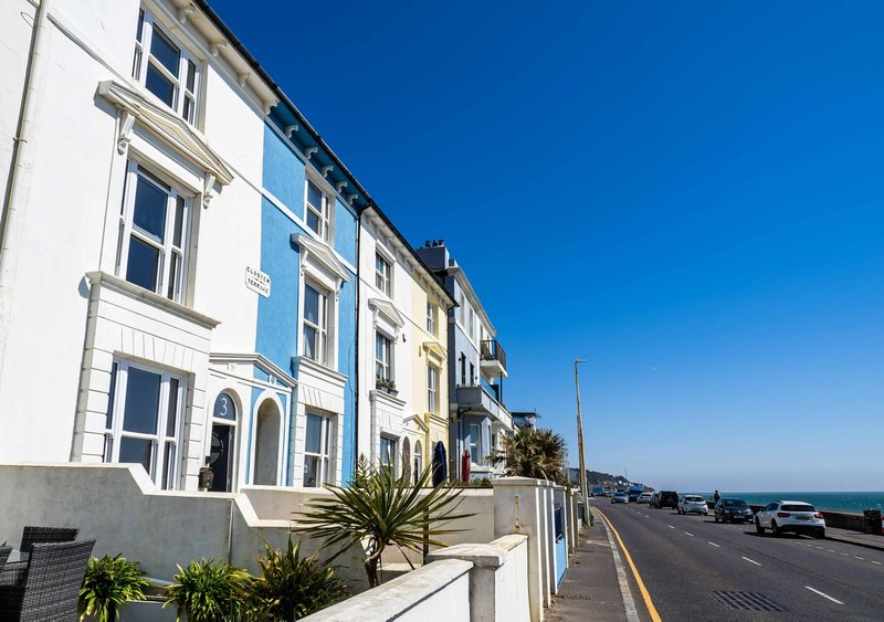 Terraced seafront building with white and blue facades, palm plants, and coastal road view