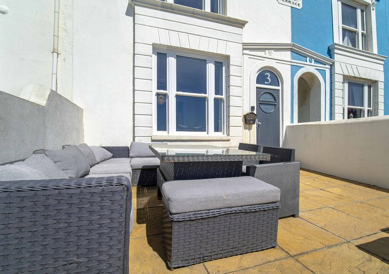 Outdoor patio seating area with grey rattan furniture and cushions, wooden decking, adjacent to colorful residential building