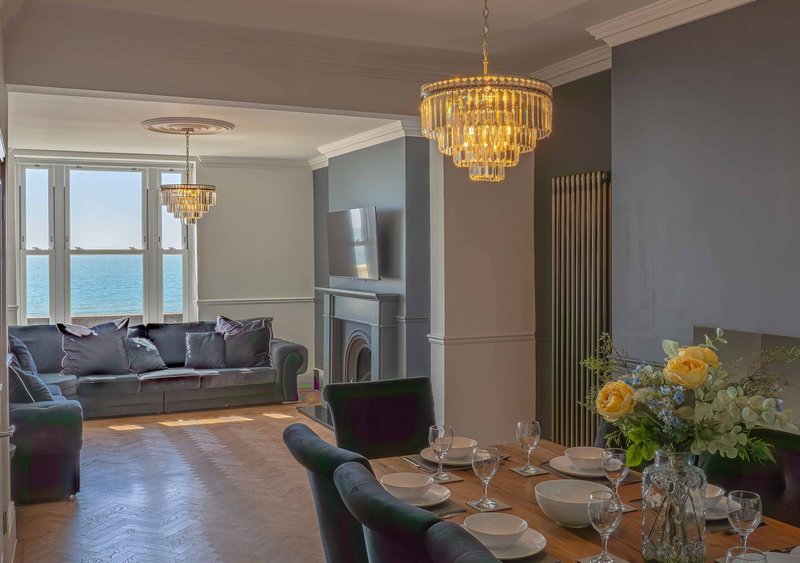 Open-plan living and dining area with sea view, dark sofa, crystal chandelier, and dining table set with flowers
