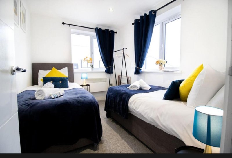 Bright bedroom with two single beds, navy bedding, yellow and blue cushions, windows with dark curtains
