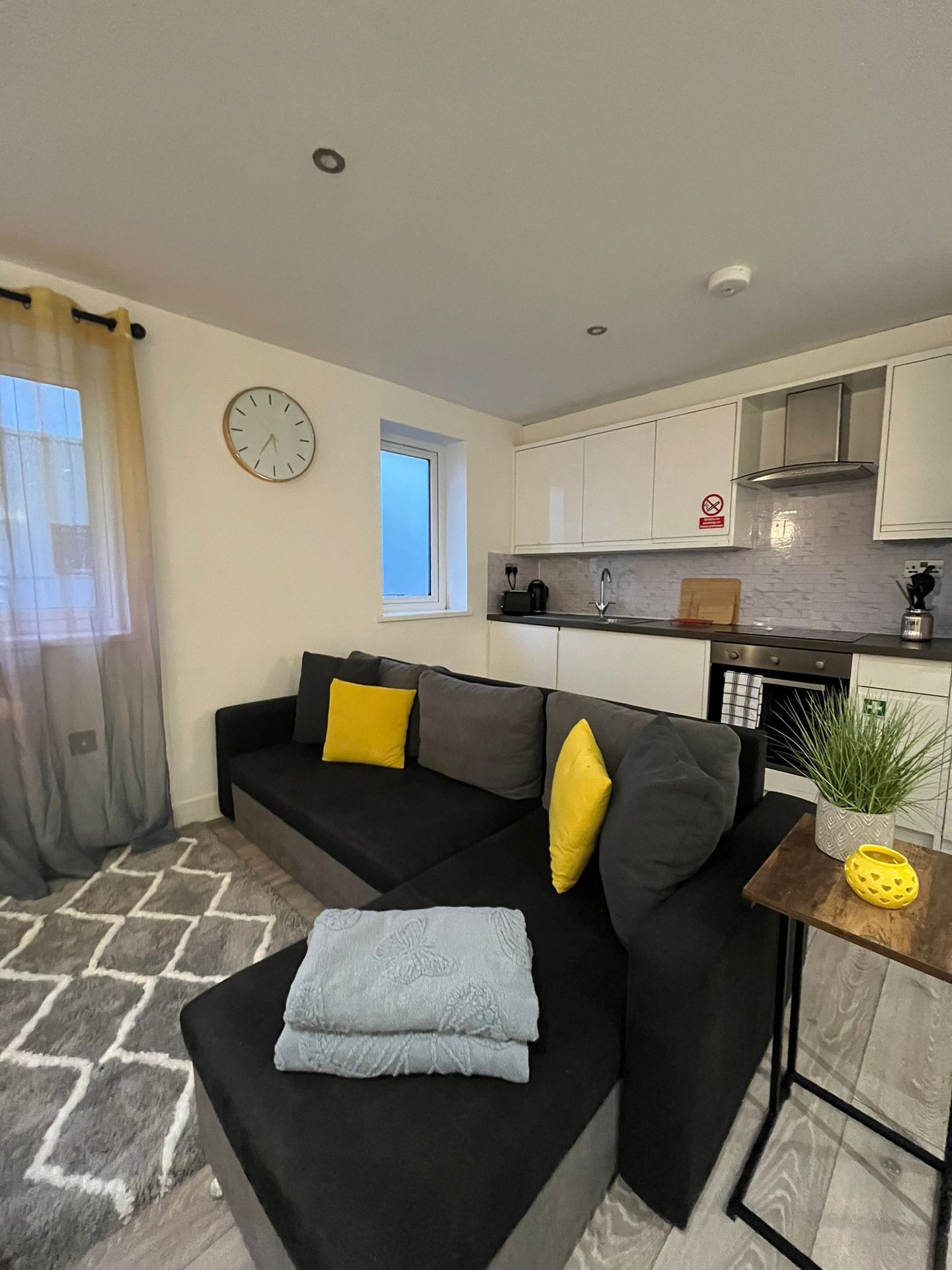 Open-plan living area with black sofa, yellow cushions, kitchenette, wall clock and windows