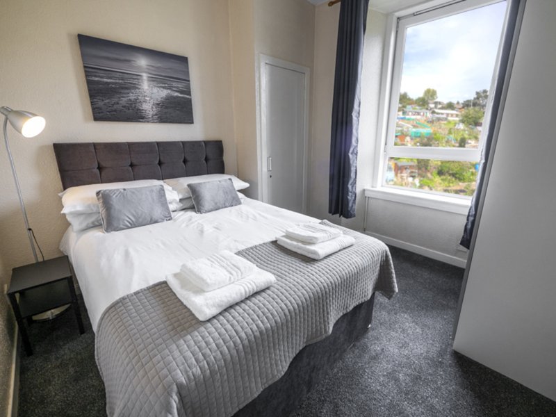 Modern bedroom with double bed, grey headboard, white bedding, and large window with garden view