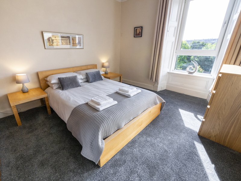 Bright bedroom with wooden bed frame, grey carpet, two bedside tables with lamps, and large window overlooking garden