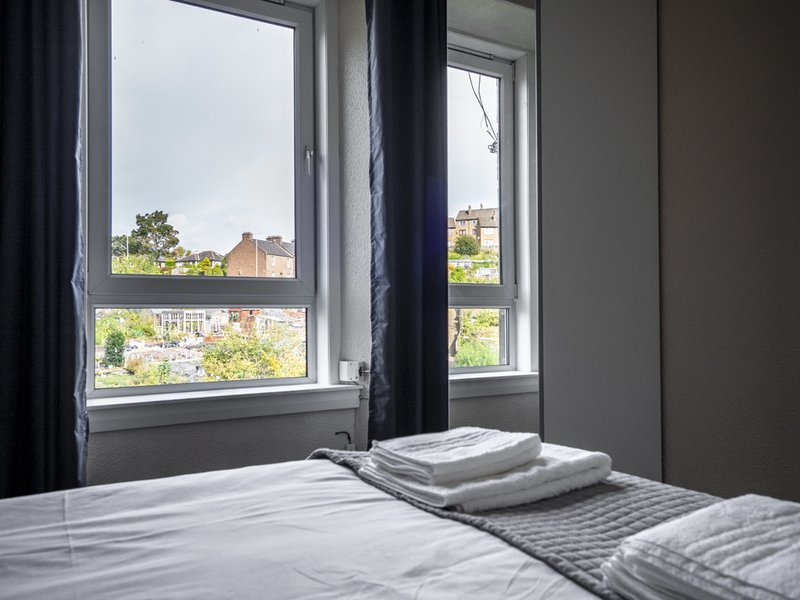 Modern bedroom with double bed, dark curtains, and windows overlooking residential area