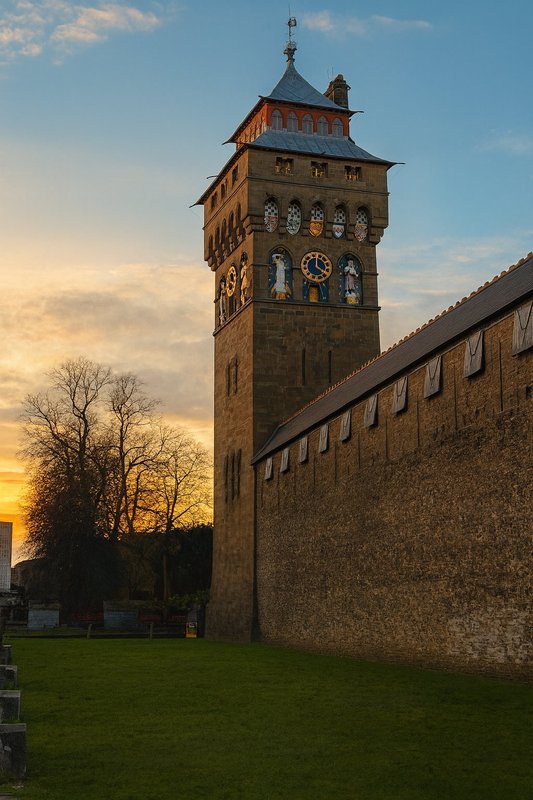 Historic stone tower with clock face at sunset, surrounded by green lawn and fortified walls