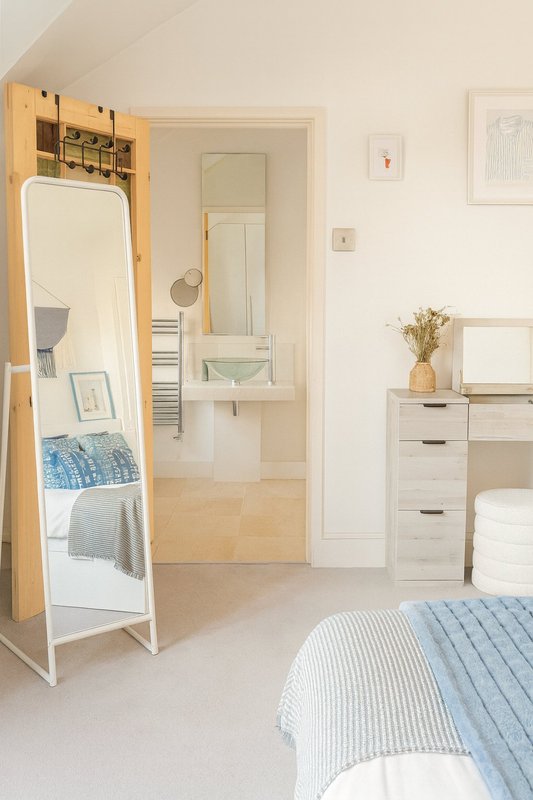 Bright bedroom with double bed, wooden shelving, mirror, dresser and ensuite bathroom visible through open door