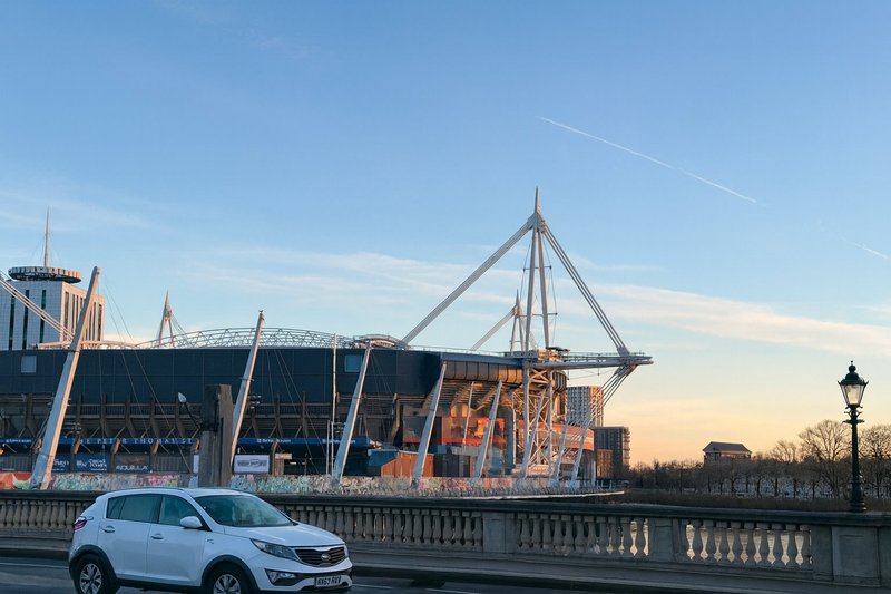 White car parked on street with large modern stadium and bridge visible across river at dusk