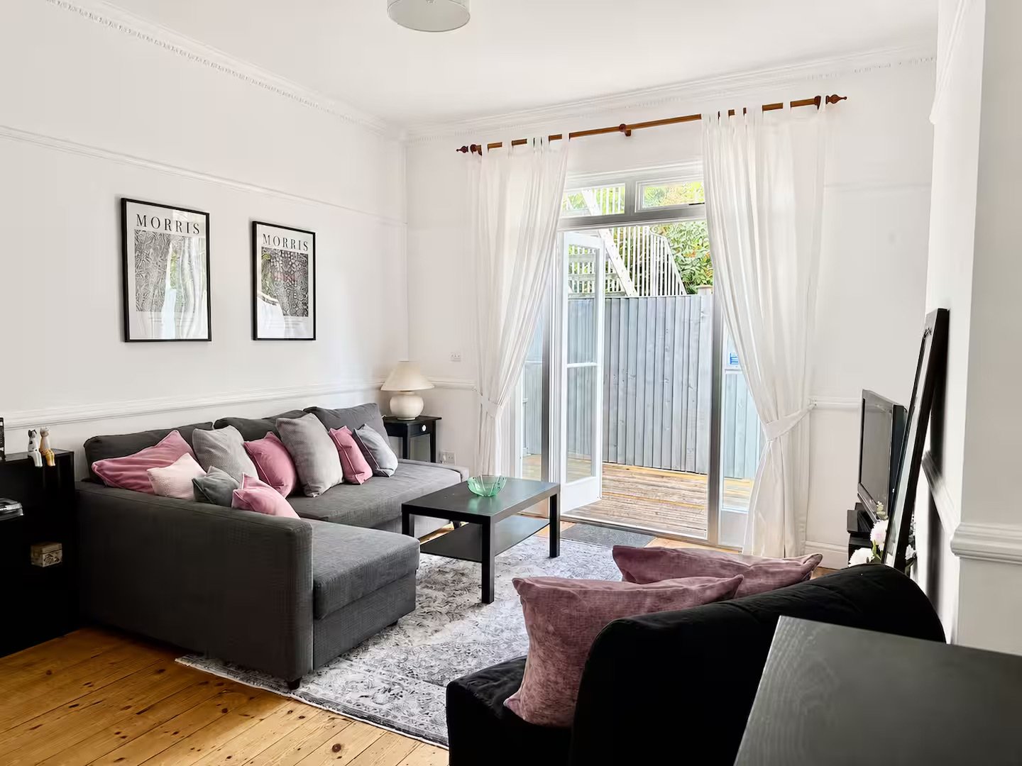 Bright living room with dark grey L-shaped sofa, pink cushions, wooden floor and glass doors to balcony