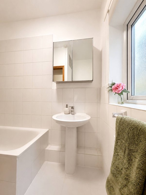 Clean white bathroom with pedestal sink, mirror, bathtub, and window with flowers