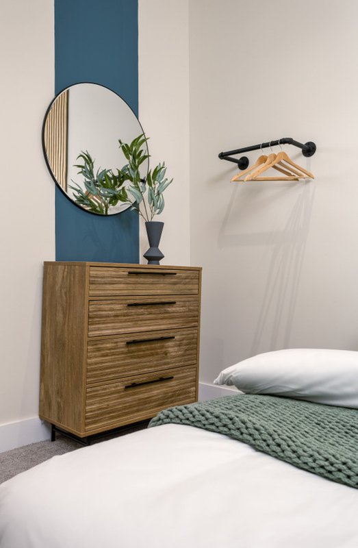Modern bedroom with wooden chest of drawers, round mirror, potted plant, and bed with white and green bedding