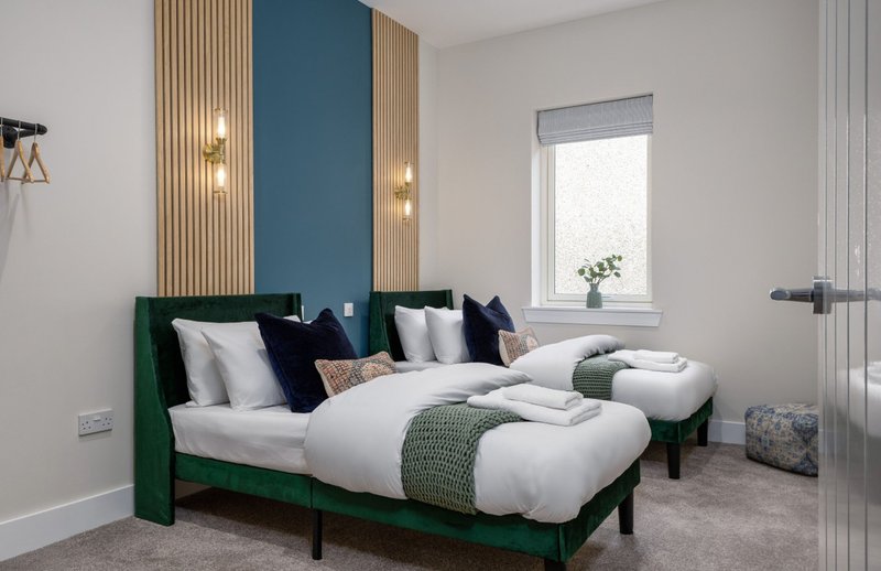 Modern bedroom with two green beds, white bedding, blue accent wall, and natural light from window