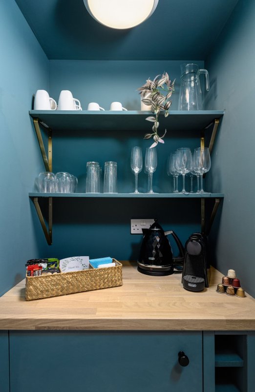 Blue kitchen alcove with open shelving displaying glassware, white bowls, flowers, kettle and small appliances