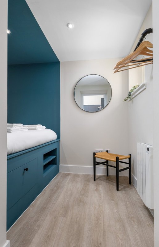 Modern bedroom nook with teal built-in bed, round mirror, wooden chair and white walls