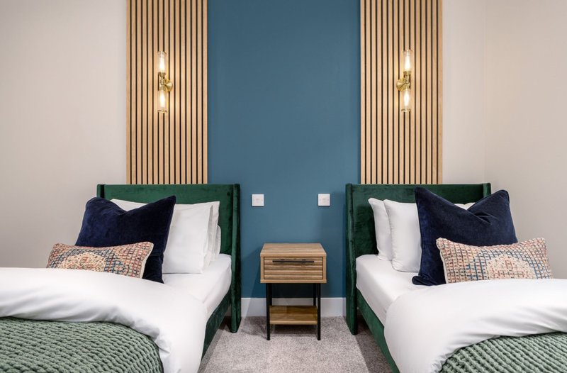 Twin bedroom with green upholstered headboards, white bedding, dark pillows, and blue accent wall