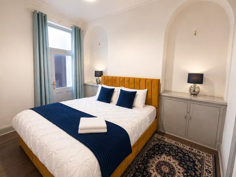 Modern bedroom with gold upholstered bed, navy bedding, alcove with built-in storage and wall-mounted TV