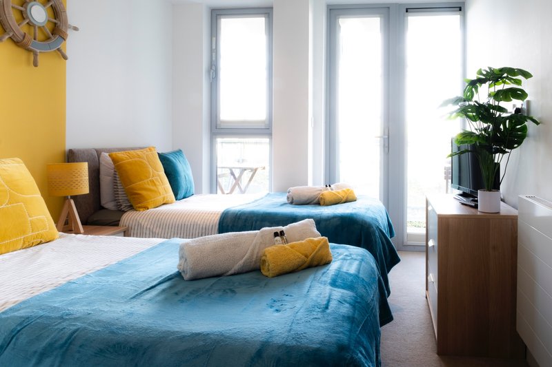 Bright bedroom with two single beds, blue bedding, yellow and teal cushions, potted plant and glass doors