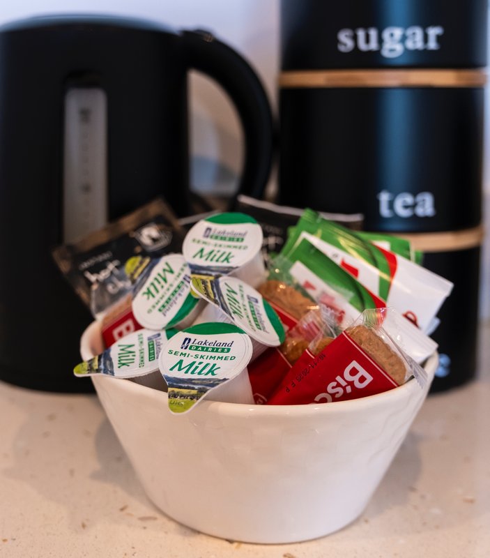 Kitchen counter with white bowl containing tea sachets, milk packets and condiments, black kettle and storage jars