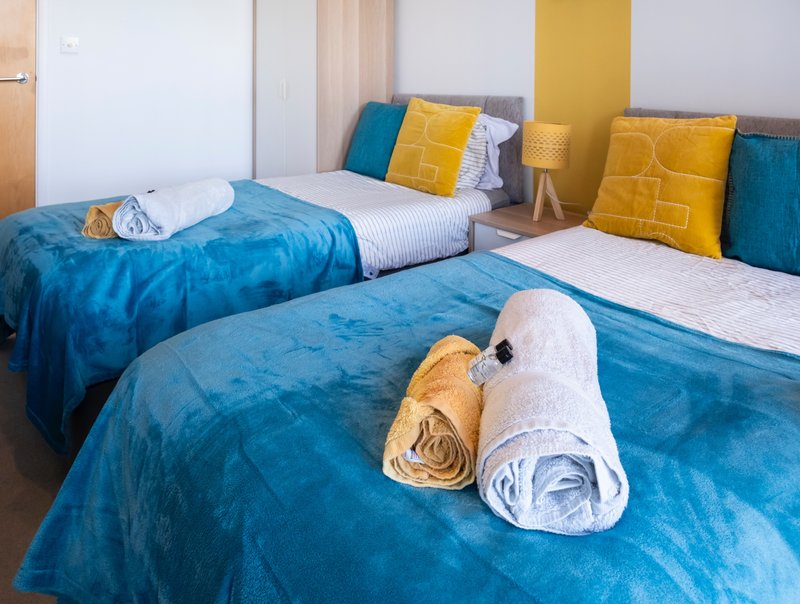Twin bedroom with blue bedding, yellow and teal cushions, rolled towels on beds