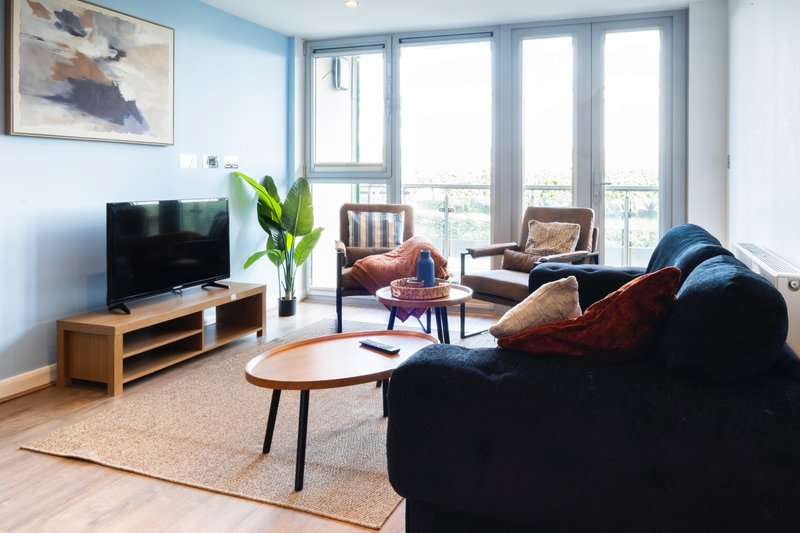 Bright modern living room with dark sofa, wooden TV unit, armchairs and large windows overlooking cityscape