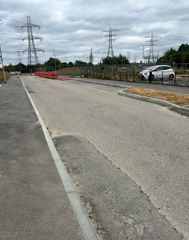 Paved driveway with parked car, fencing, and electricity pylons in background