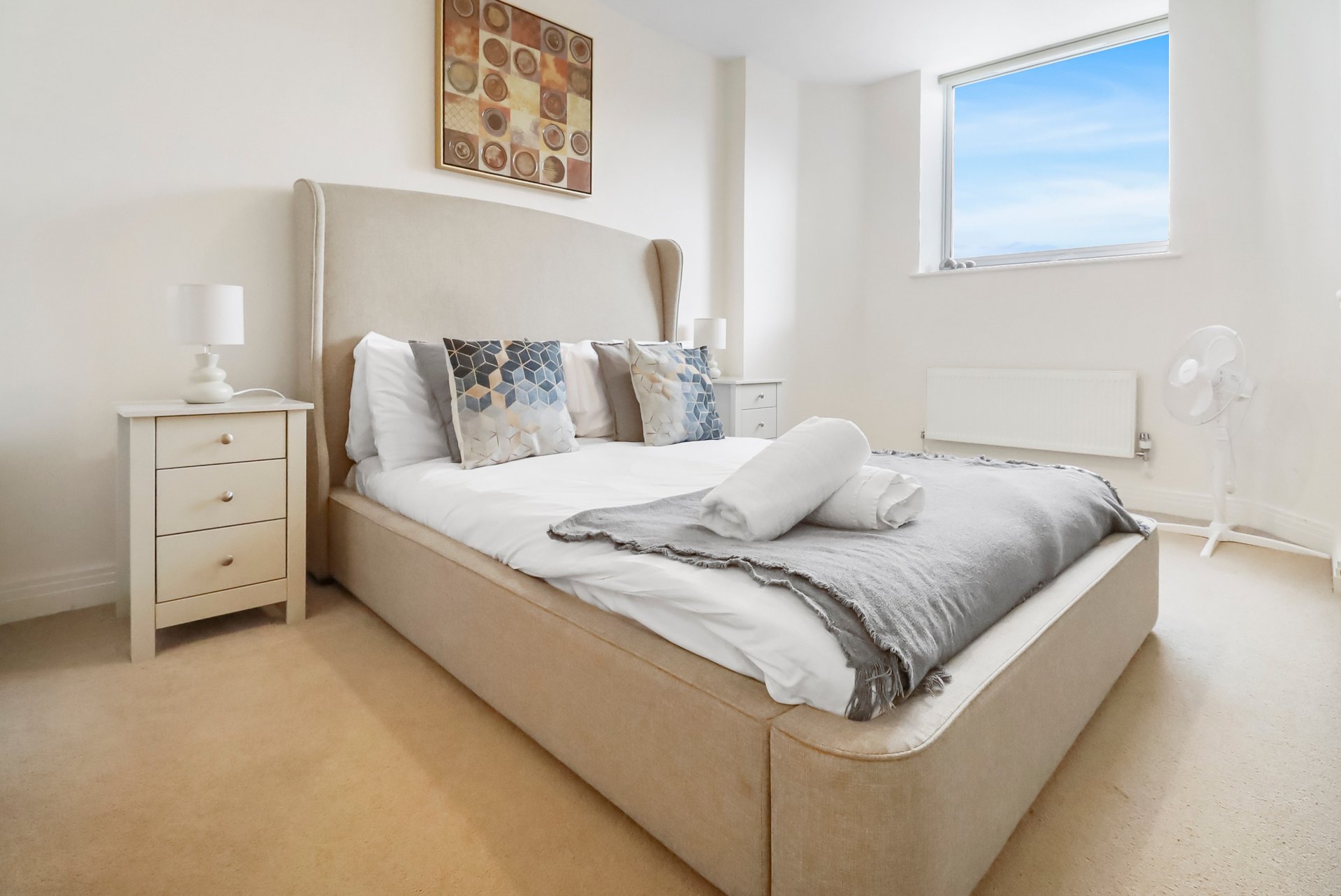Bright modern bedroom with upholstered bed, bedside table, geometric wall art and window with blue sky view