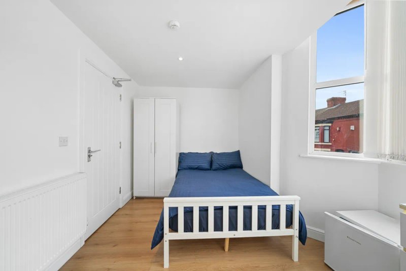 Bright bedroom with white walls, single bed with blue bedding, wooden floor, and street view window