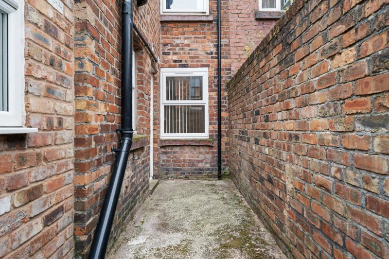 Narrow brick alleyway between two properties with central window and downpipe