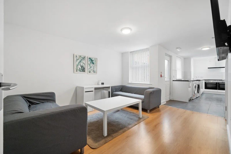 Open-plan living area with grey sofa, white coffee table, kitchen appliances and wooden flooring