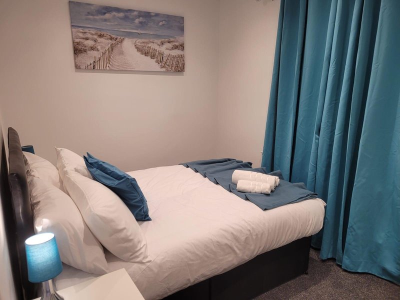 Bedroom with double bed, white bedding, blue cushions, framed landscape artwork, and teal curtains