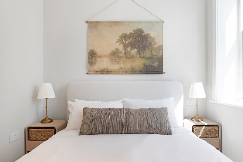 Bright bedroom with white bed, gray cushion, framed landscape art, and wooden bedside tables with lamps