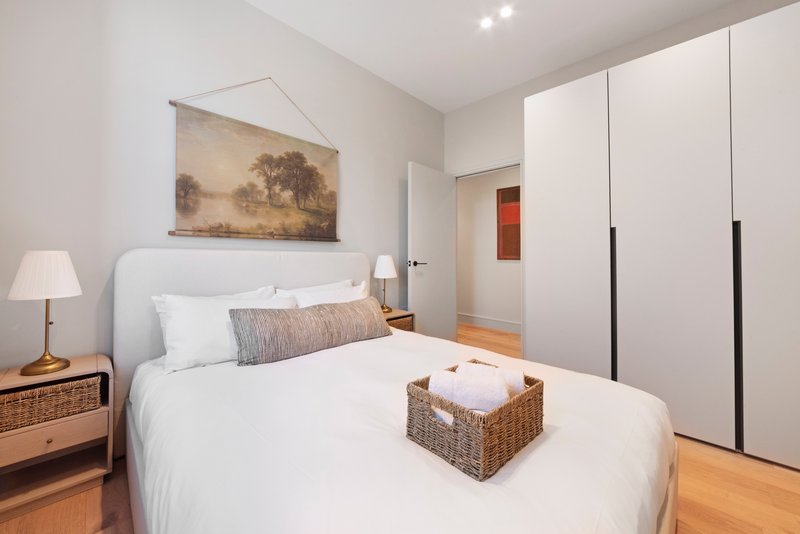 Modern bedroom with white bed, wooden nightstands, framed landscape art, and white wardrobe