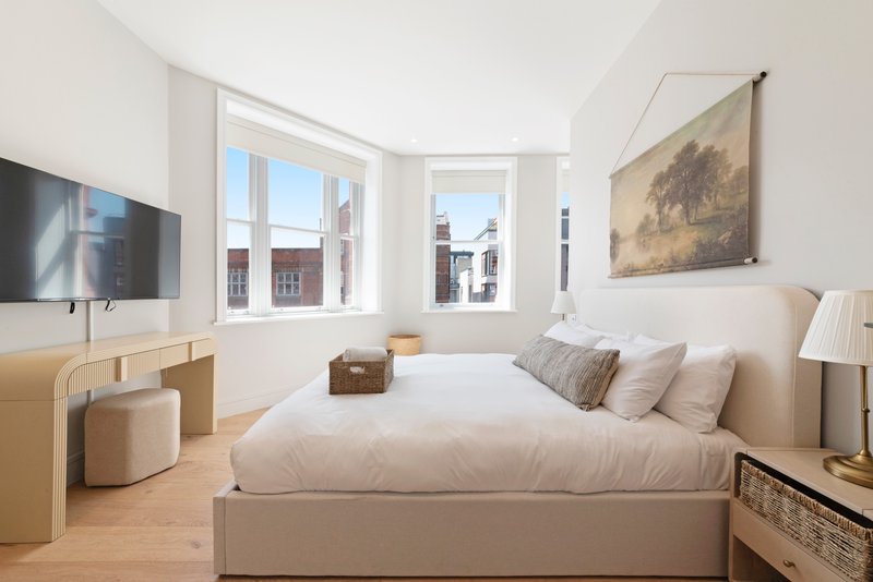 Bright modern bedroom with double bed, wooden furniture, wall-mounted TV and city views through windows