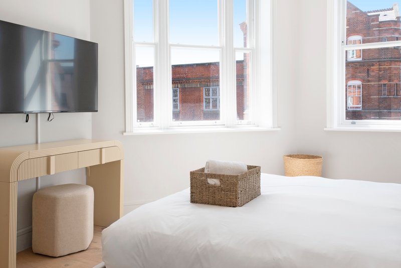 Bright bedroom with double bed, wooden desk, wall-mounted TV and large windows overlooking brick buildings
